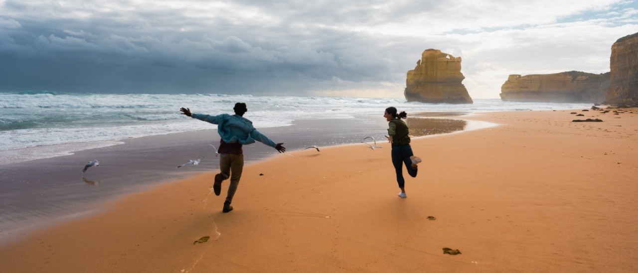 two people running down a beach with the 12 Apostles in background