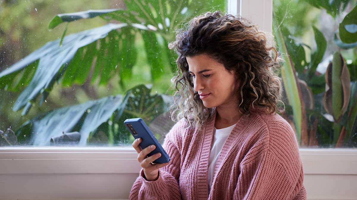 Woman in pink cardigan making mobile phone call in room with large ferns outside window behind