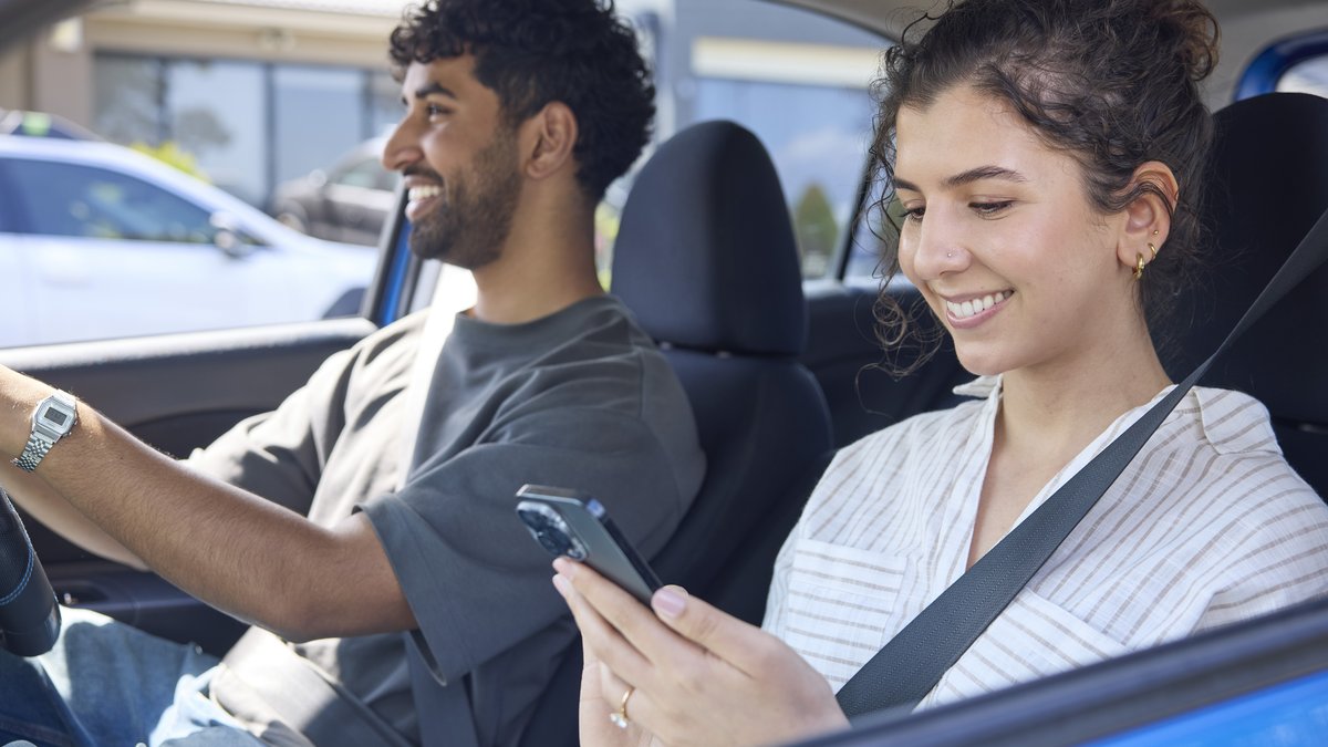 A man drives happily as a woman in the passenger seat uses the RACV App on her phone