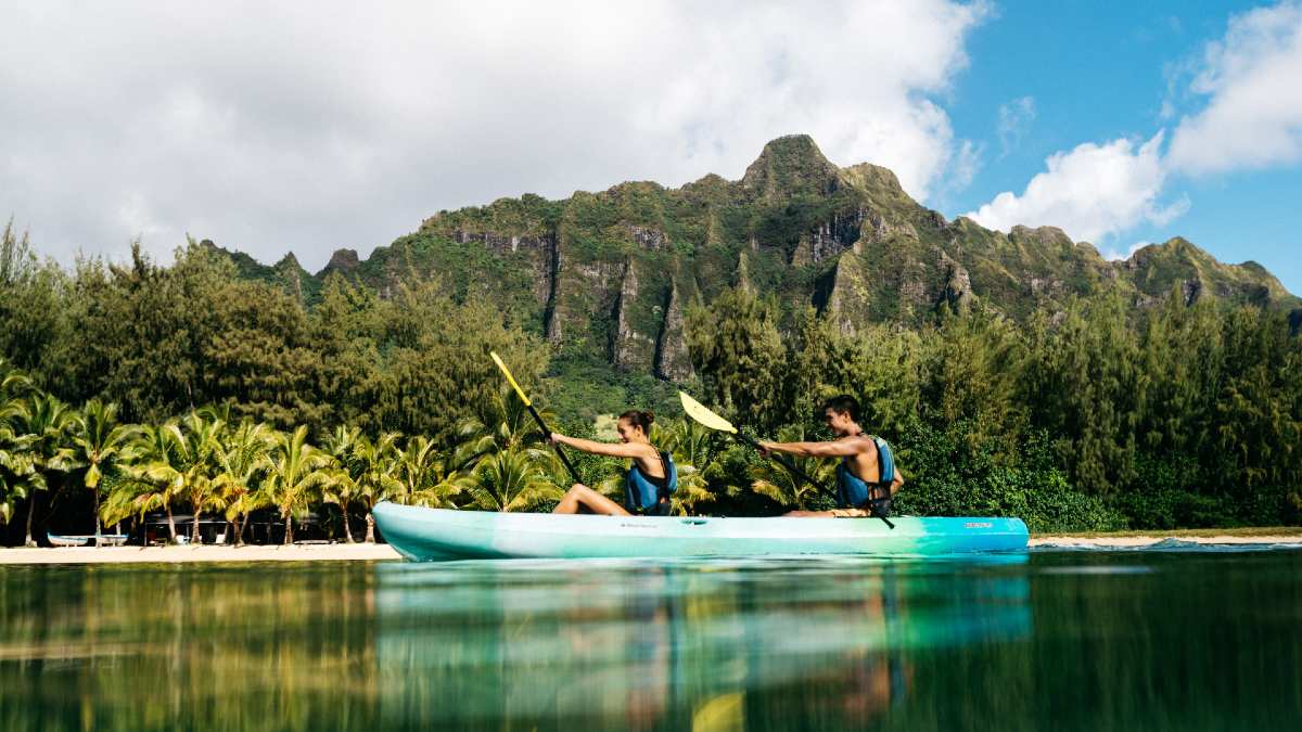 two people kayaking in ocean near shore lined with lush forest and mountains