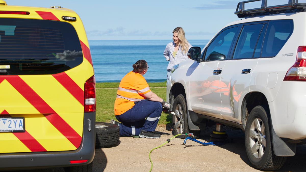 An RACV emergency roadside assistance technician helping fix a flat tyre on a white SUV while chatting to the owner of the car