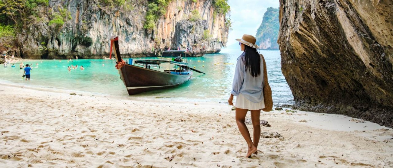 woman walking on a sandy Thai beach with boats, swimmers and islands in background