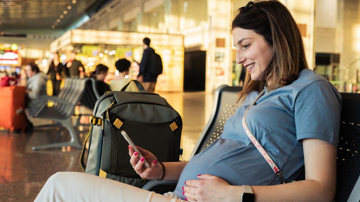 pregnant woman touching her stomach in an airport seat, smiling at her phone