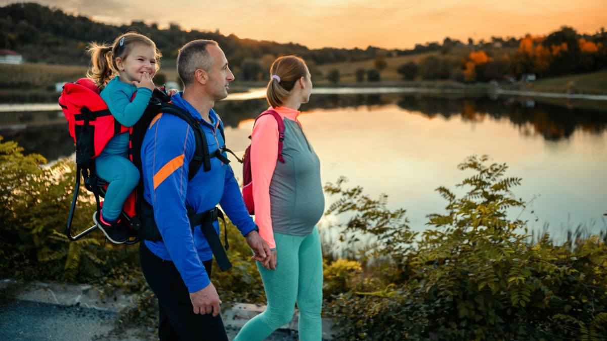man carrying child on back walking next to pregnant woman near a lake