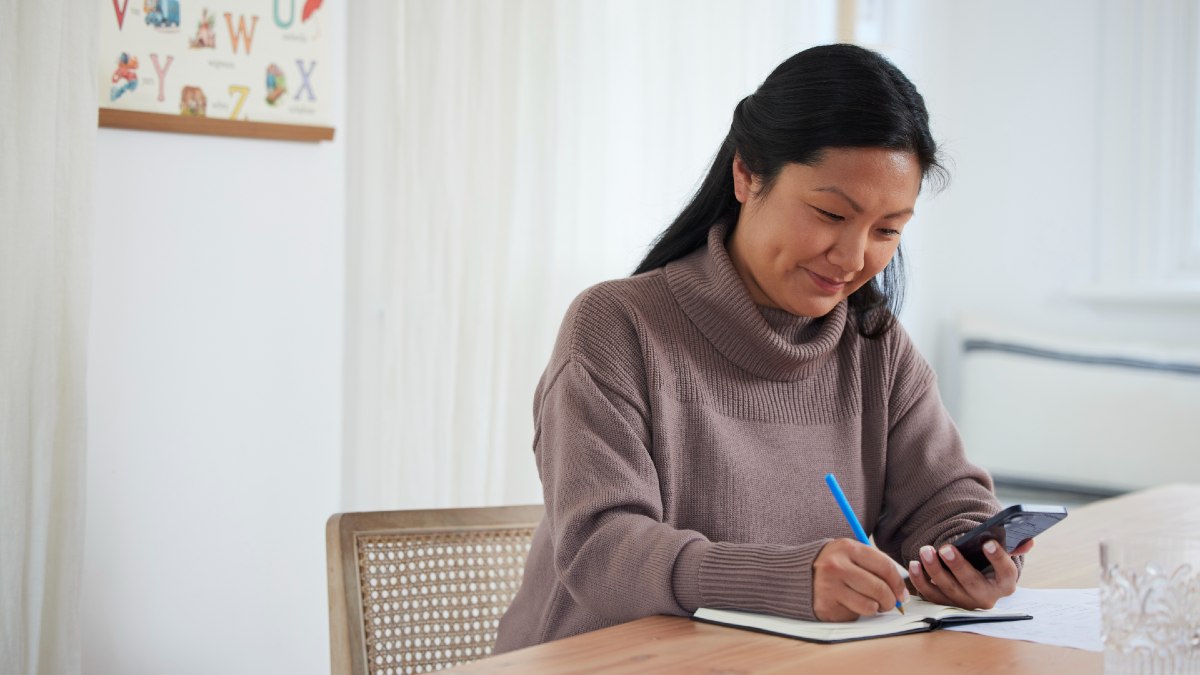 woman looking at smartphone and smiling while writing in a notepad