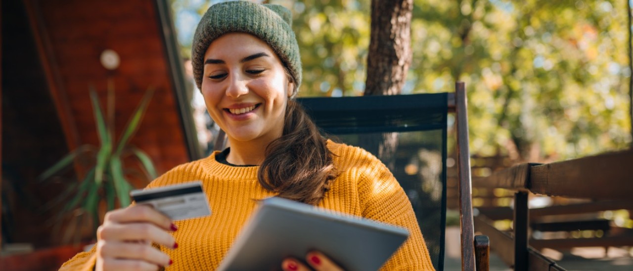 young woman in yellow sweater and green beanie sitting on a deck smiling at a credit card, holding a tablet