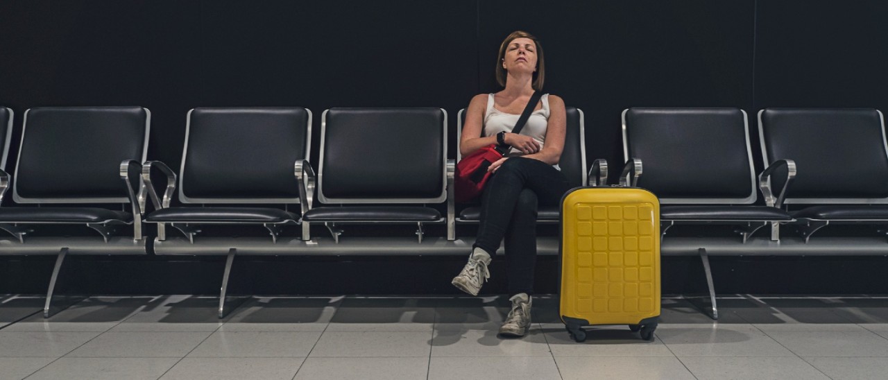 A tired-looking woman sitting on a row of seats waiting for her flight at an airport. She leans back on her chair with her eyes closed and legs crossed while a bright yellow suitcase is on the ground next to her