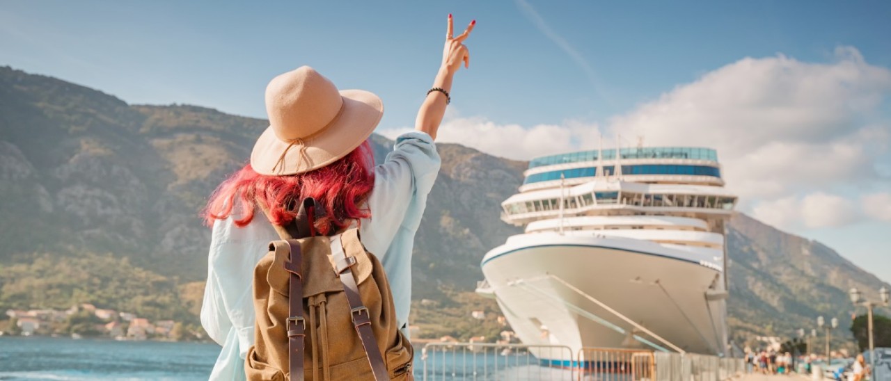 woman with red hair, sunhat and backpack giving a peace sign to a large cruise ship
