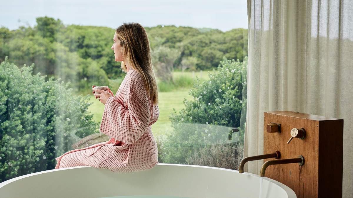 woman in pink robe sitting on edge of bathtub with cup of tea, looking over gardens