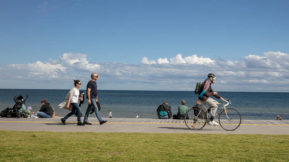 Older person riding bike on St Kilda foreshore shared pathway with pedestrians sitting on boardwalk behind