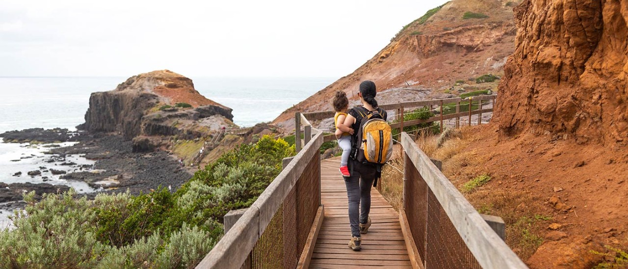 Mother carrying toddler on boardwalk towards Pulpit Rock at Cape Schanck on the Mornington Peninsula.