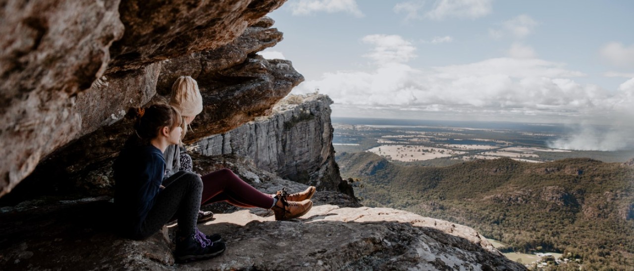 Two people sitting on a rock ledge in the GRampians and overlooking a spectacular vista of the land below