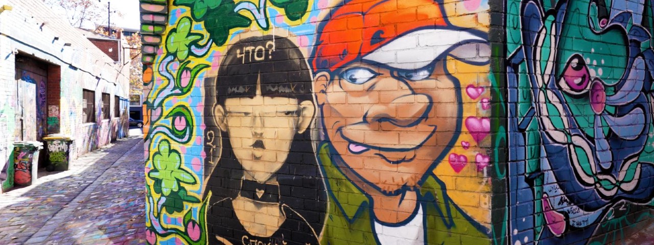 A colourful street art mural featuring a goth-looking woman and a goofy-looking man wearing a baseball cap in a Melbourne laneway