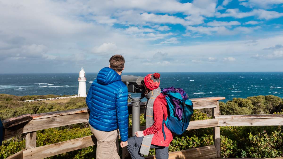 young man and woman looking through telescope at a white lighthouse by the ocean