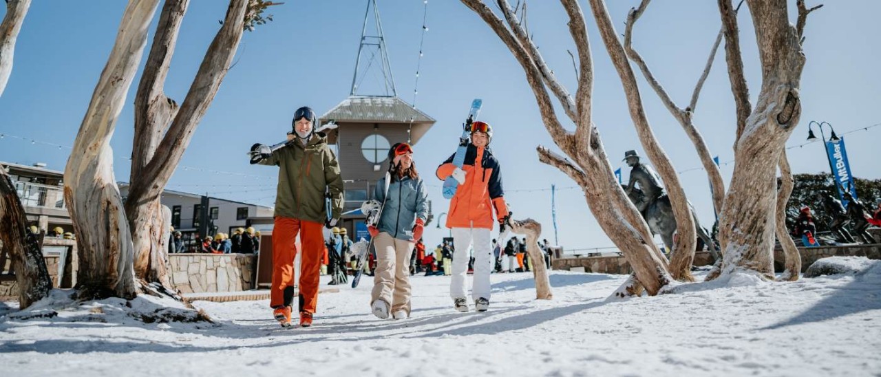 Skiing at Mt Buller_Visit Victoria three friends dressed in snow gear carrying skis at Mt Buller, Victoria
